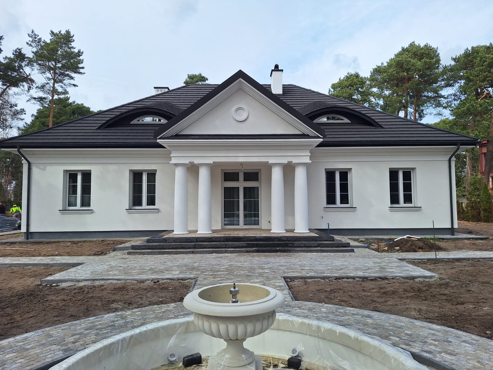 Elegancki, biały dom jednorodzinny z kolumnami i ciemnym dachem, otoczony zielenią. Na pierwszym planie fontanna z kamienia.