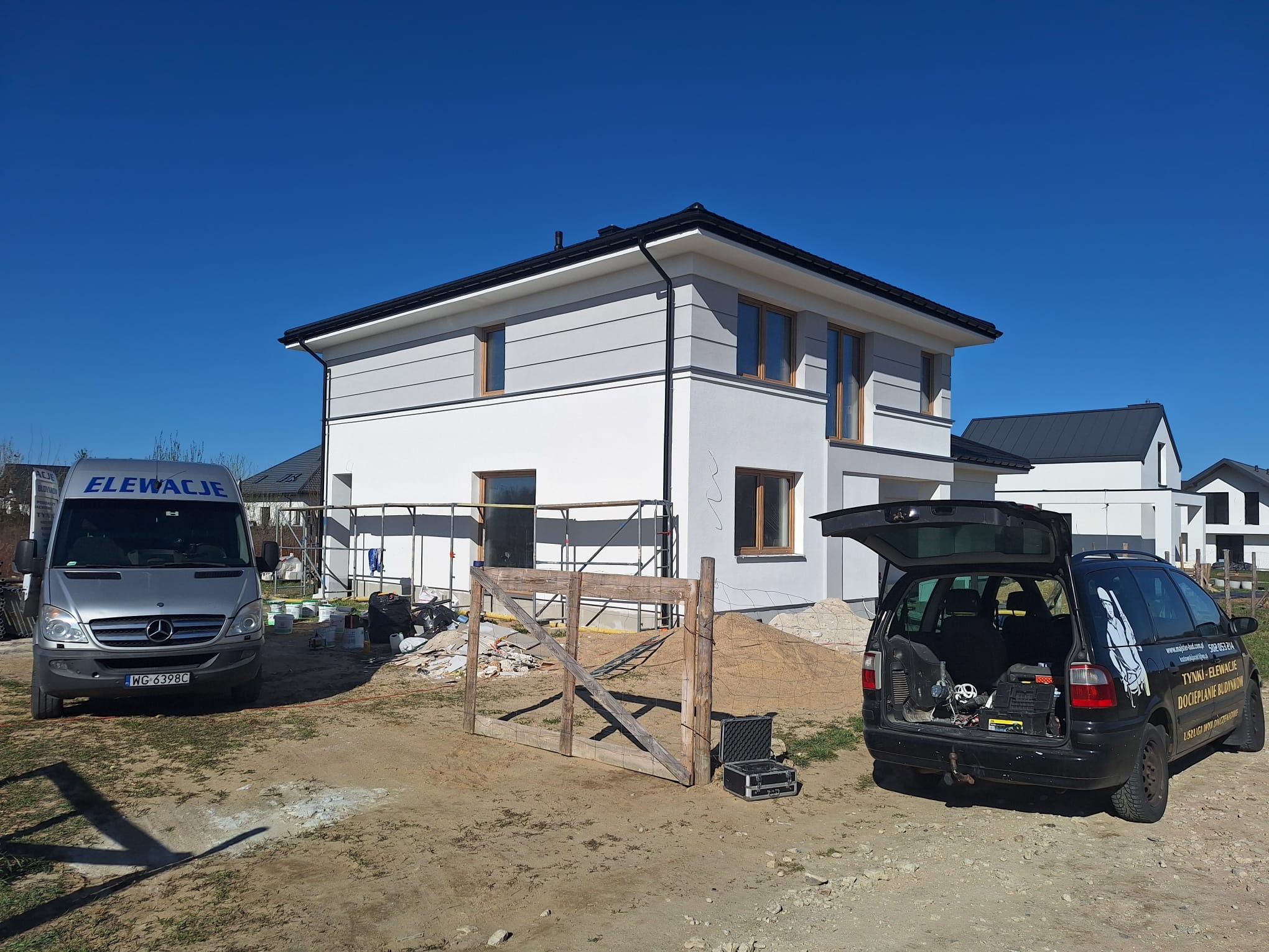 Dwukondygnacyjny dom z szarą i białą elewacją, częściowo ocieplony. Na pierwszym planie samochód z reklamą firmy elewacyjnej i rusztowanie. Błękitne niebo.