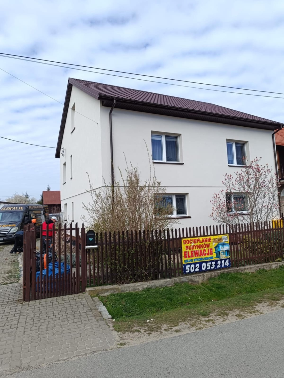 Dwupiętrowy dom z jasną elewacją i brązowym dachem, widoczny zza drewnianego płotu z reklamą usług ocieplania. Częściowo widoczny bus i pracownicy.
