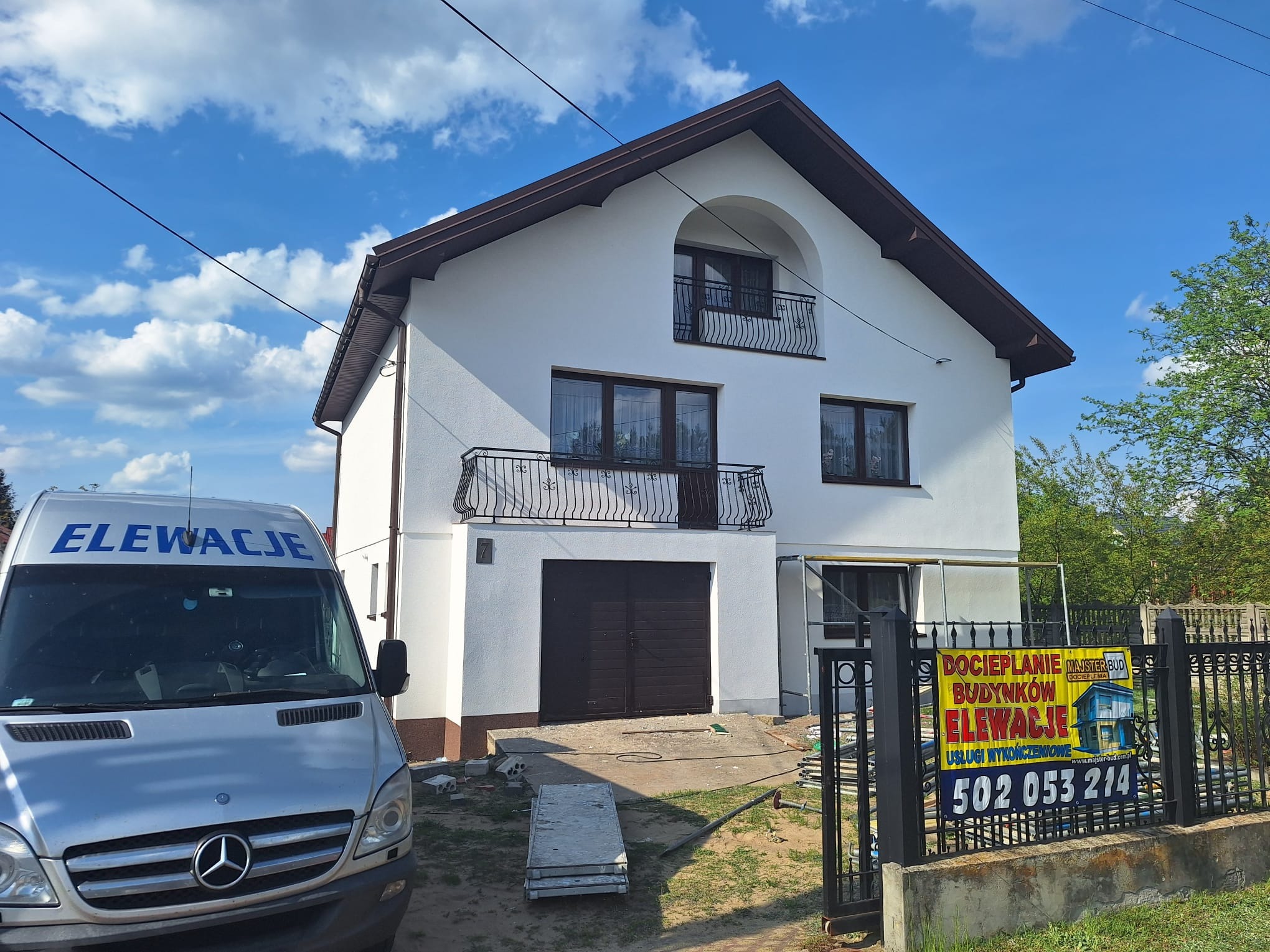 Biały dom z brązowymi akcentami po renowacji elewacji, widoczny samochód z napisem 'Elewacje' i baner reklamowy firmy. Słoneczny dzień.