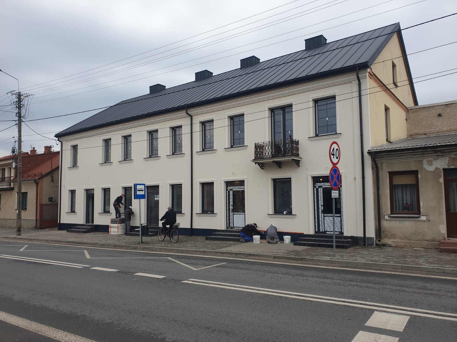 Pracownicy przy białej elewacji budynku, jeden na drabinie, inni na ziemi. Widoczny rowerzysta i znak przystanku. Czarny dach i okna. Ulica z liniami.