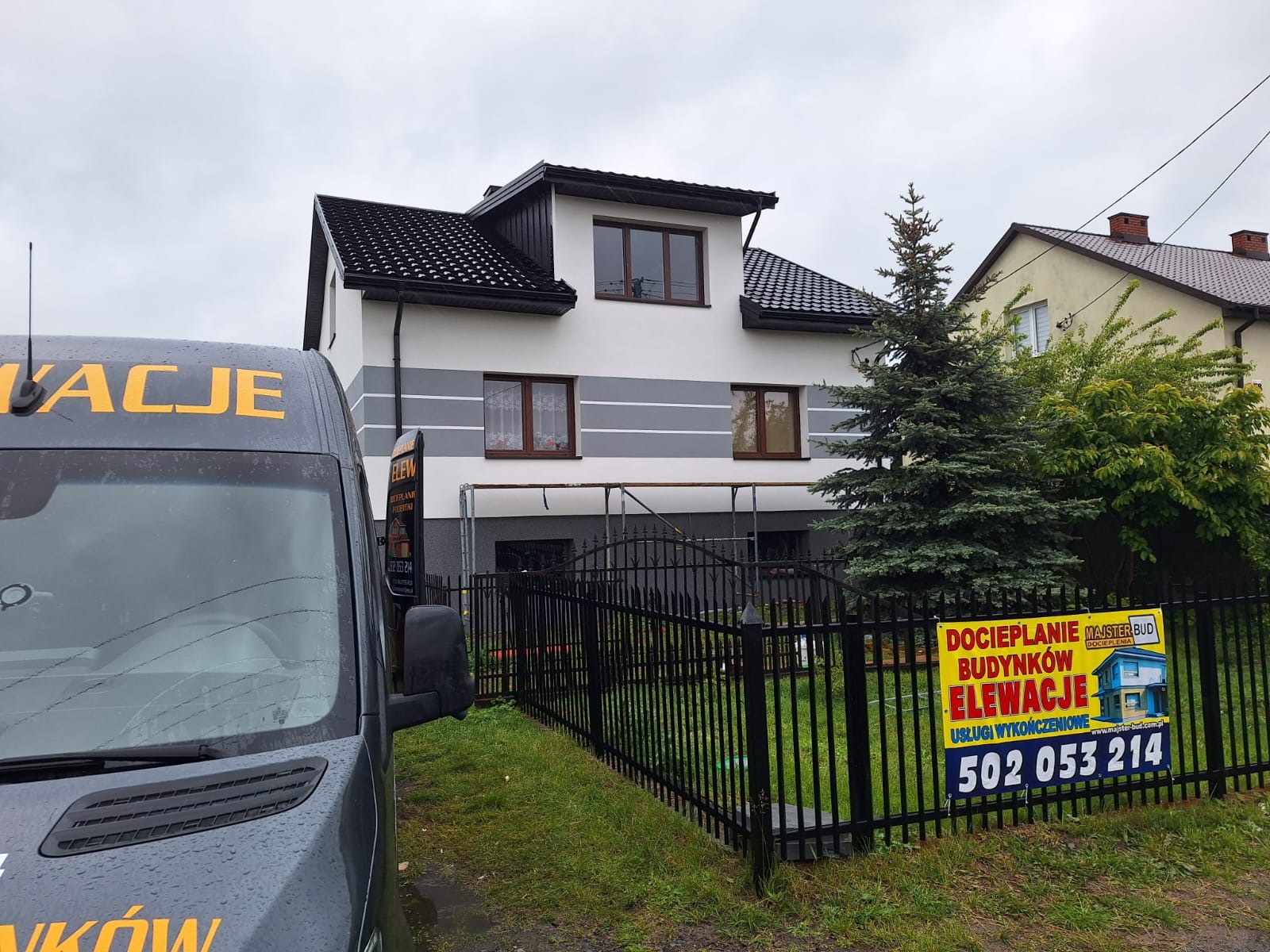Dom po renowacji elewacji z szarymi pasami, widoczny zza ogrodzenia i częściowo zasłonięty przez samochód z reklamą firmy. Na ogrodzeniu baner reklamowy.