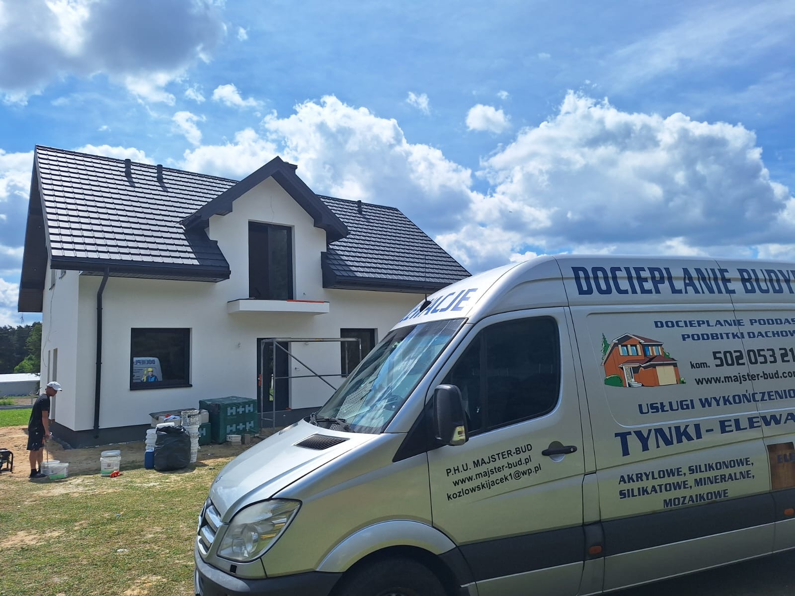 Nowoczesny dom z białą elewacją i czarnym dachem, widoczny zza srebrnego busa z reklamą firmy budowlanej. Na trawniku stoją puszki z farbą i pracownik.