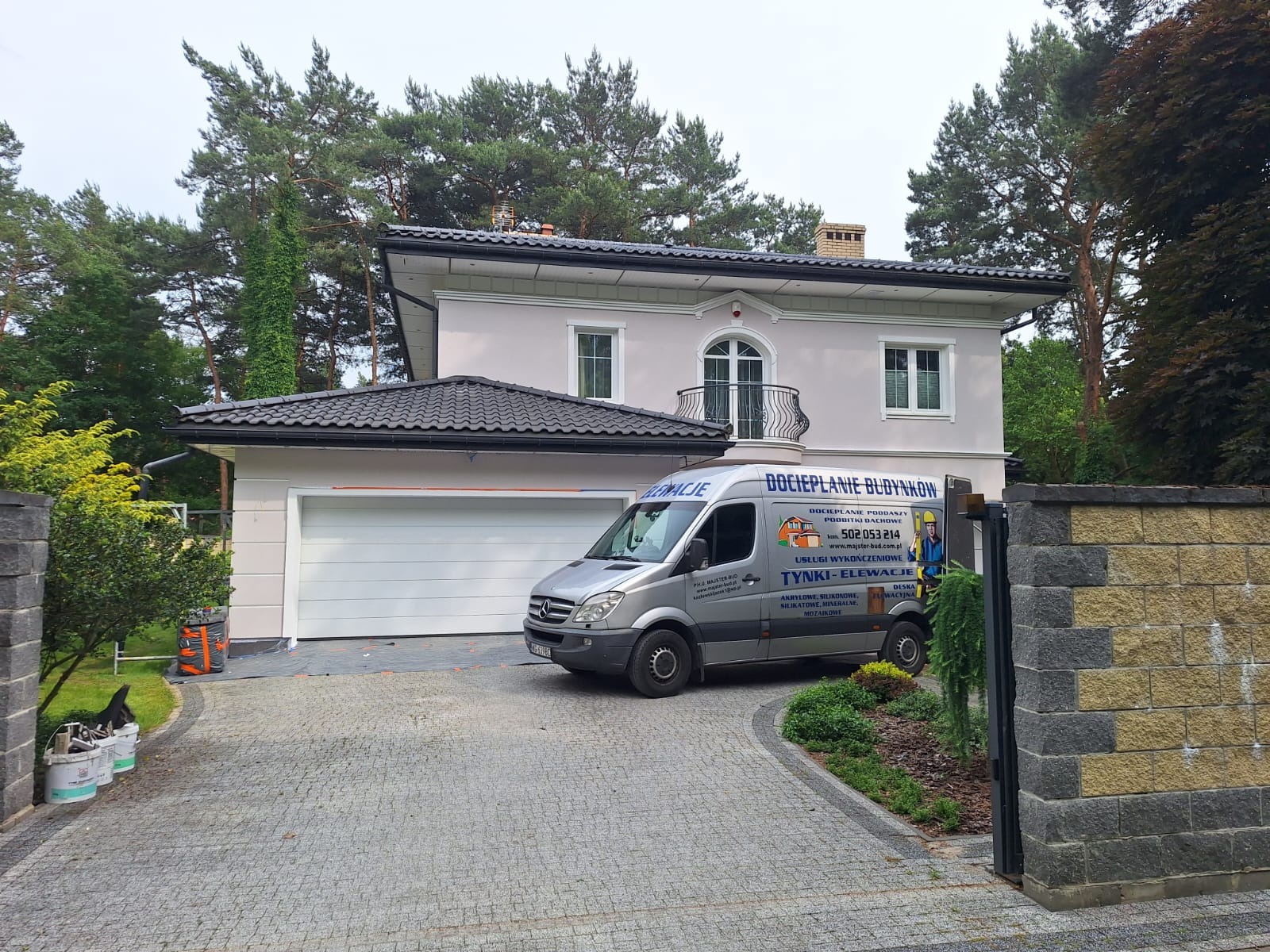 Jasny, dwukondygnacyjny dom z garażem i balkonem, po renowacji elewacji. Na podjeździe zaparkowany srebrny bus z reklamą usług budowlanych. Widok z zewnątrz, w tle drzewa.