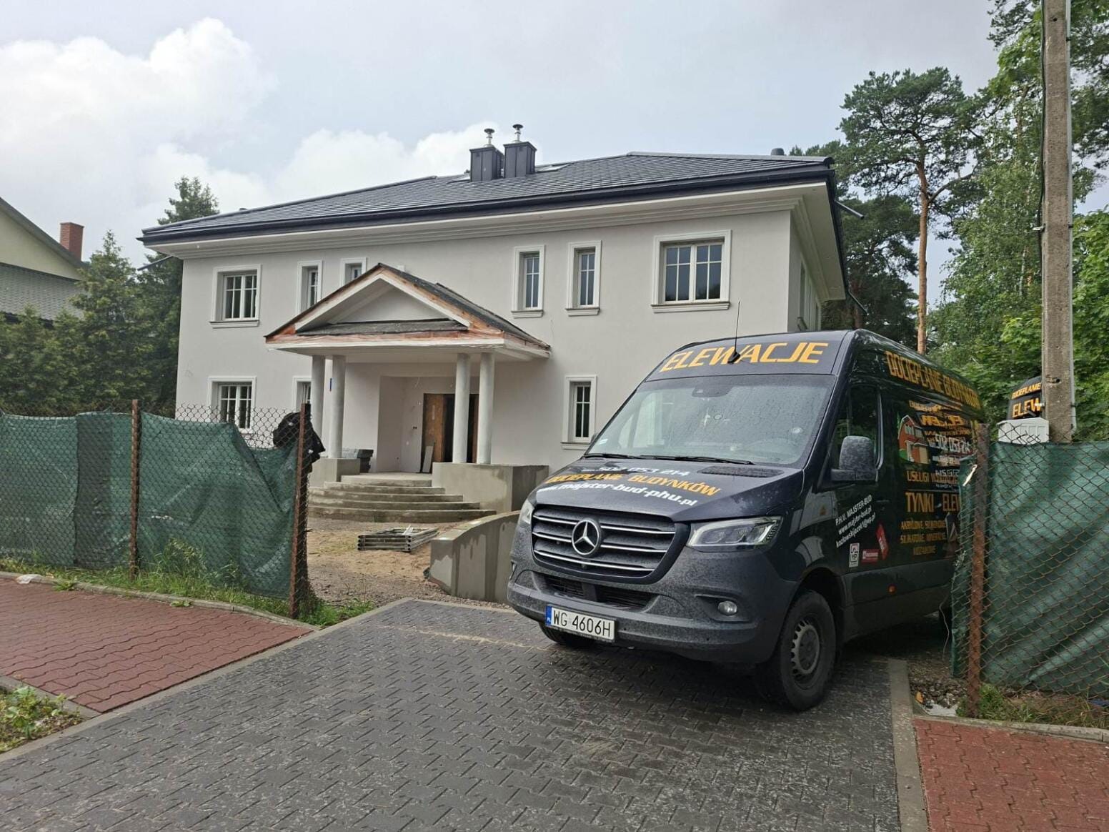 Jasny, dwukondygnacyjny dom z szarą elewacją i ciemnym dachem, częściowo zasłonięty przez firmowy bus z napisem 'Elewacje' i zielone ogrodzenie.