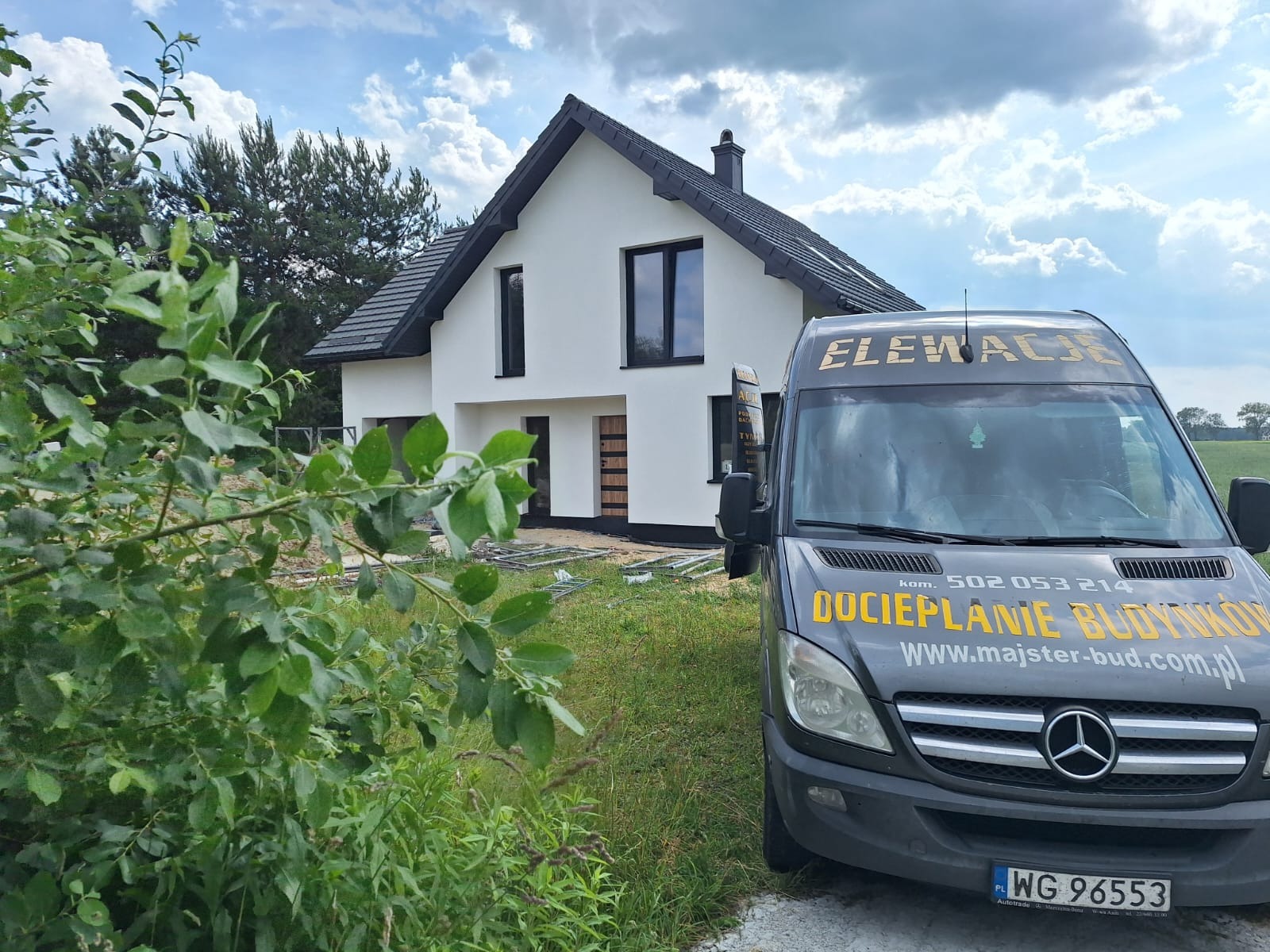 Nowoczesny dom z białą elewacją i czarną stolarką okienną, na pierwszym planie firmowy bus z napisem 'Elewacje' i 'Ocieplanie Budynków'.