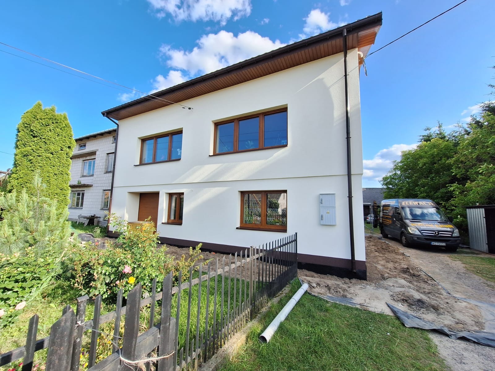 Dwukondygnacyjny dom po renowacji elewacji, z brązowymi oknami i dachem, na tle błękitnego nieba z chmurami. Widoczny ogród i samochód dostawczy z napisem 'Elewacje'.