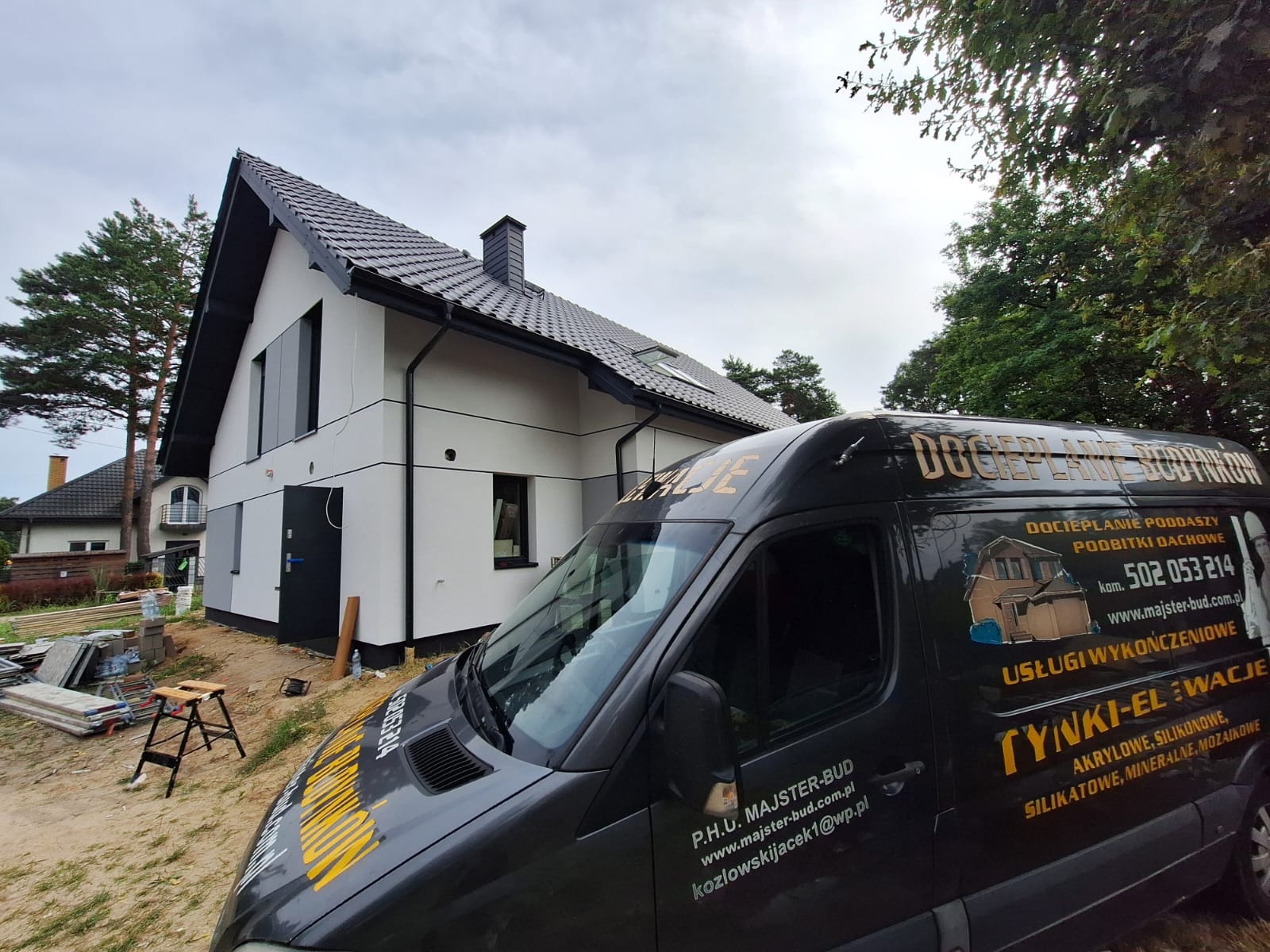 Dom po renowacji elewacji w Sulbinach, widoczny zza firmowego busa z reklamą usług: ocieplanie poddaszy, tynki elewacyjne. Nowoczesny wygląd budynku.