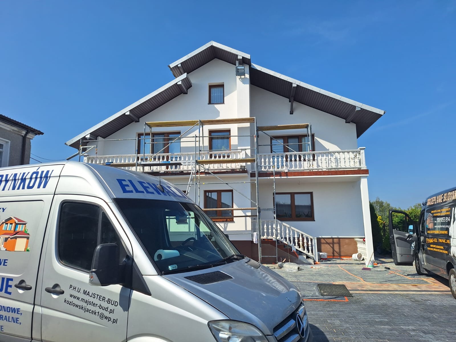 Dwupiętrowy dom z balkonem w trakcie prac elewacyjnych, widoczne rusztowania. Na pierwszym planie samochód dostawczy z reklamą firmy budowlanej.