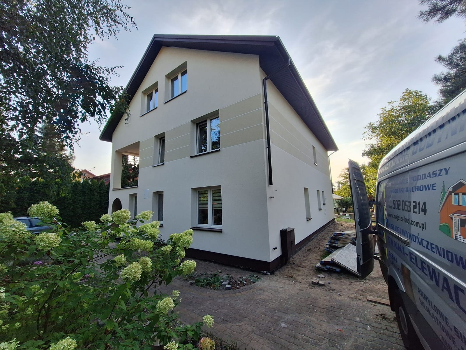 Dwukondygnacyjny dom z jasną elewacją, z pasami w odcieniach beżu. Widoczny fragment busa z reklamą firmy budowlanej. Ujęcie z poziomu ogrodu, w tle zieleń.
