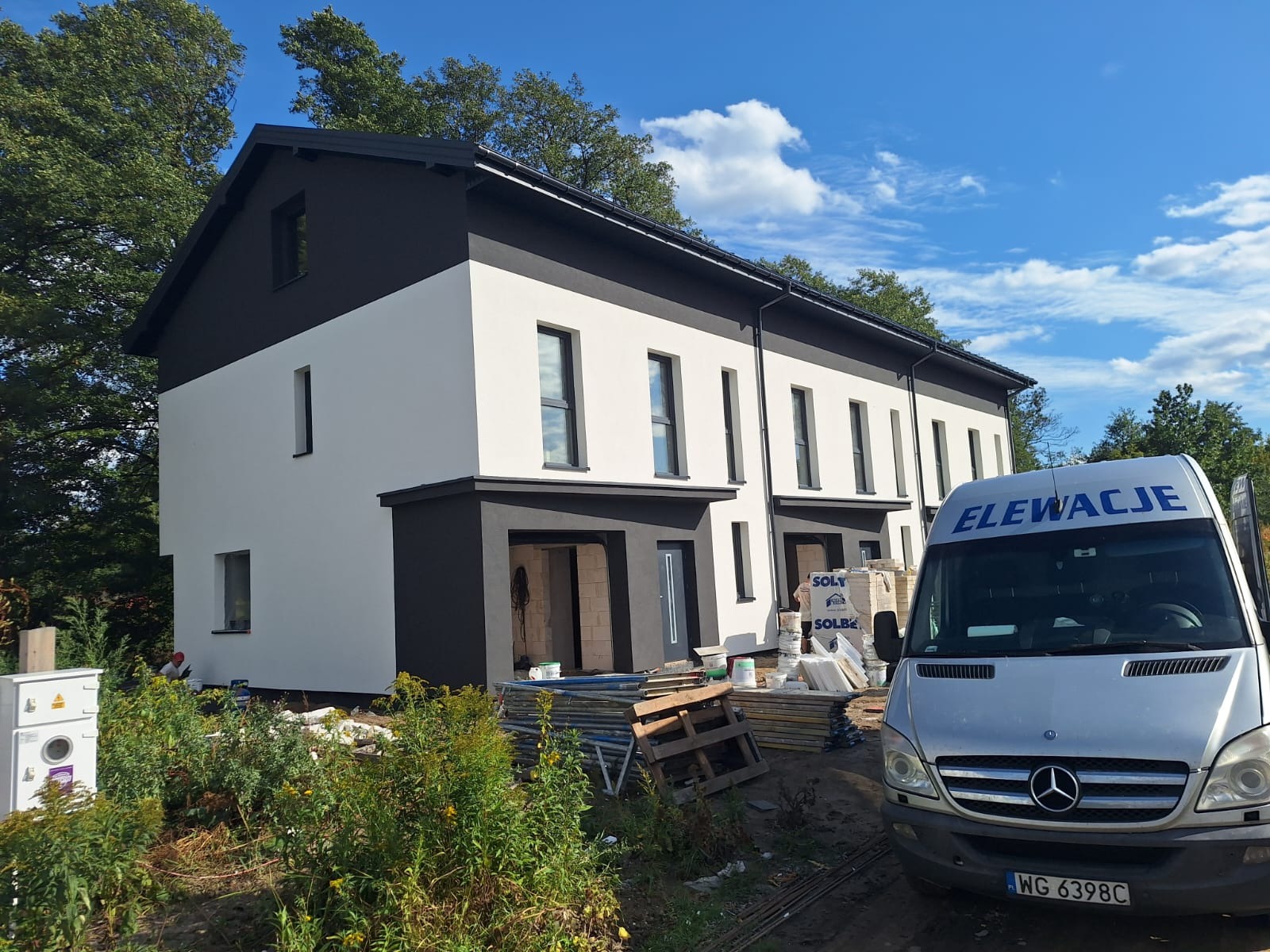 Nowoczesny dom dwukondygnacyjny z biało-czarną elewacją, widoczny z boku, z samochodem dostawczym z napisem 'Elewacje' na pierwszym planie i materiałami budowlanymi.