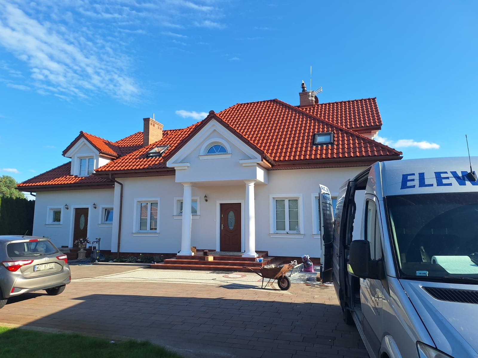 Jasny dom jednorodzinny z czerwoną dachówką, widoczny samochód osobowy i dostawczy z napisem 'ELEW', taczka i akcesoria malarskie. Błękitne niebo z chmurami.