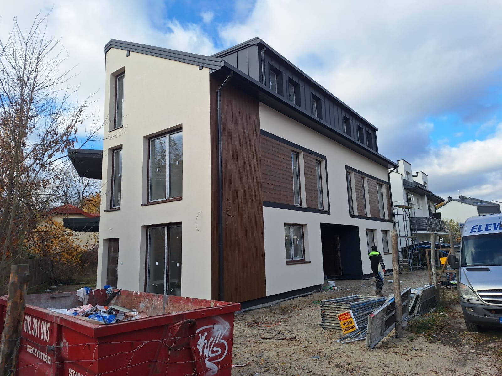 Nowoczesny budynek po renowacji elewacji z elementami drewnianymi, widoczny kontener na gruz i pracownik w tle. Jasna kolorystyka i minimalistyczny design.