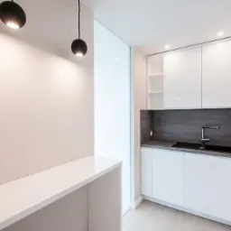 Nowoczesna, minimalistyczna kuchnia z białymi błyszczącymi szafkami, blatem z szarego kamienia i czarnym zlewem, oświetlona kulistymi lampami wiszącymi na tle kremowej ściany.