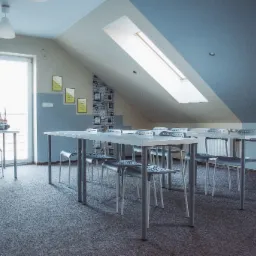 Jasna sala lekcyjna na poddaszu z białymi stołami, szarymi krzesłami i oknem dachowym, ściana z dekoracyjną tapetą imitującą cegły z napisami.