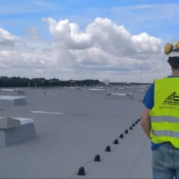 Pracownik w żółtej kamizelce z logo firmy na dachu budynku przemysłowego podczas montażu instalacji odgromowej. Widoczne uziomy i przewody odgromowe. W tle pochmurne niebo.