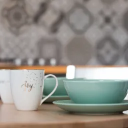 Zestaw śniadaniowy: biały kubek z napisem 'joy' i złotymi gwiazdkami obok miętowej miseczki na talerzyku, na drewnianym blacie, z rozmytym tłem kuchennym w szarościach.