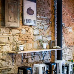 Surowa ściana z cegły i kamienia z plakatem Wielkopolski, drewniana półka z dekoracjami, czarne i białe stołki barowe w kawiarni.