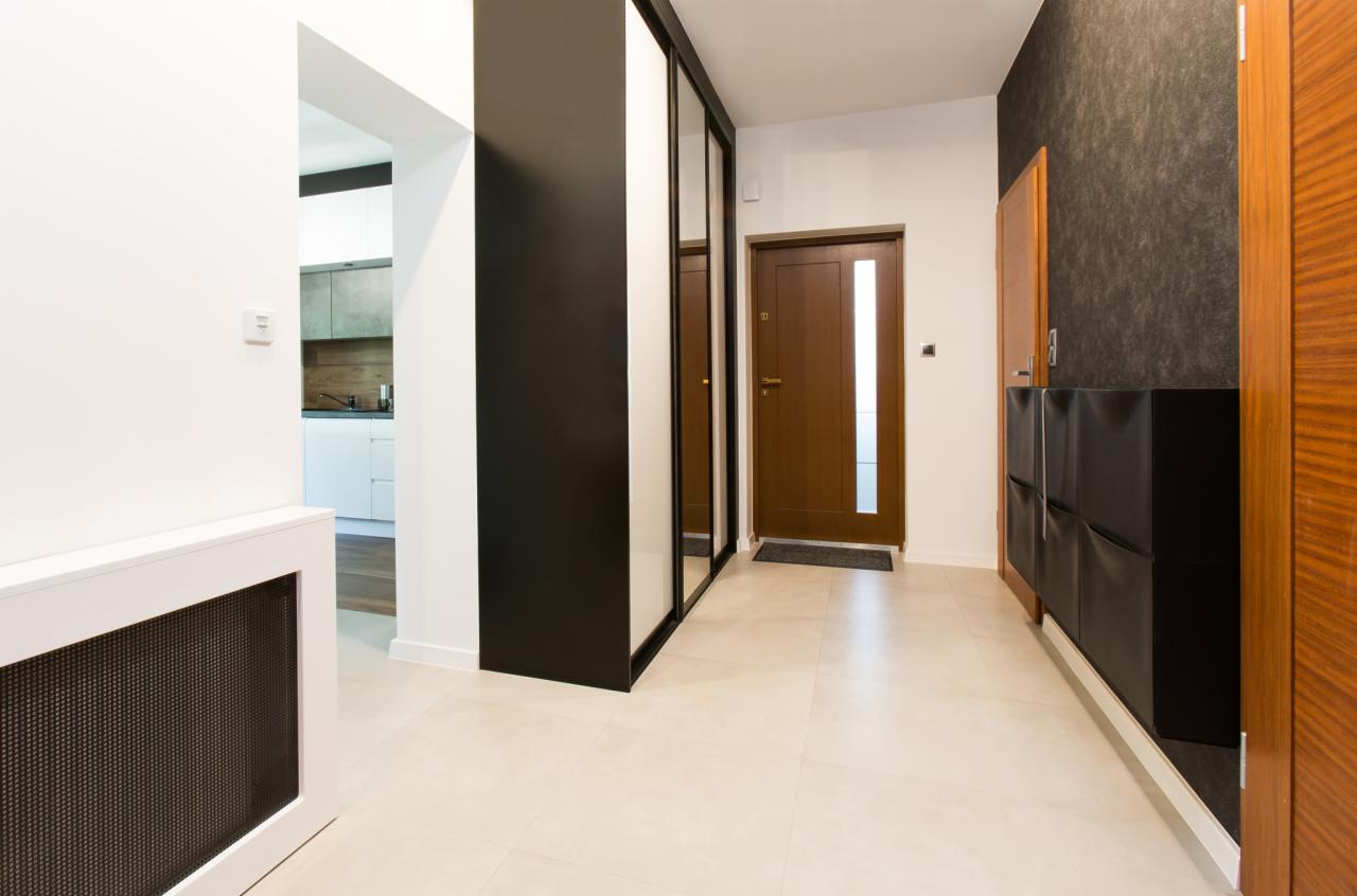 Nowoczesny przedpokój z beżową podłogą, czarną szafą z lustrami, drewnianymi drzwiami wejściowymi z szybą i czarnym organizerem na buty.