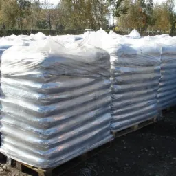 Rzędy palet z workami opału owiniętymi folią, składowane na zewnątrz, widoczne drzewa w tle.