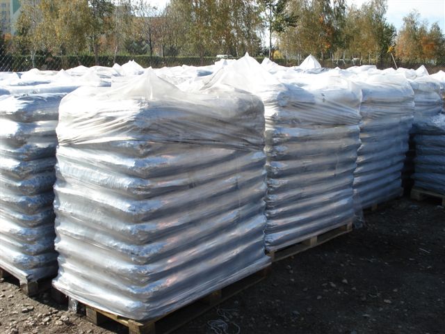 Rzędy palet z workami opału owiniętymi folią, składowane na zewnątrz, widoczne drzewa w tle.