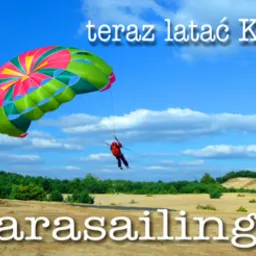 Osoba w czerwonym kombinezonie uprawiająca parasailing na tle piaszczystego terenu z lasem i błękitnego nieba z chmurami. Na dole zdjęcia widoczny adres strony internetowej.