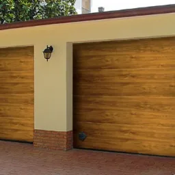 Dwie zamknięte, segmentowe bramy garażowe o fakturze drewna, umieszczone w jasnym budynku z ceglanymi akcentami, oświetlone zewnętrzną lampą.