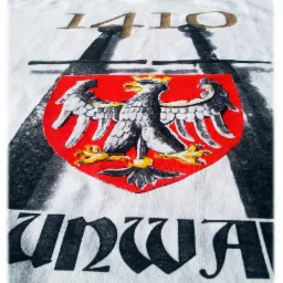Biała koszulka z nadrukiem herbu z orłem na czerwonym tle, daty 1410 i napisu 'Grunwald' w gotyckim stylu. Widok z bliska, lekko z góry.