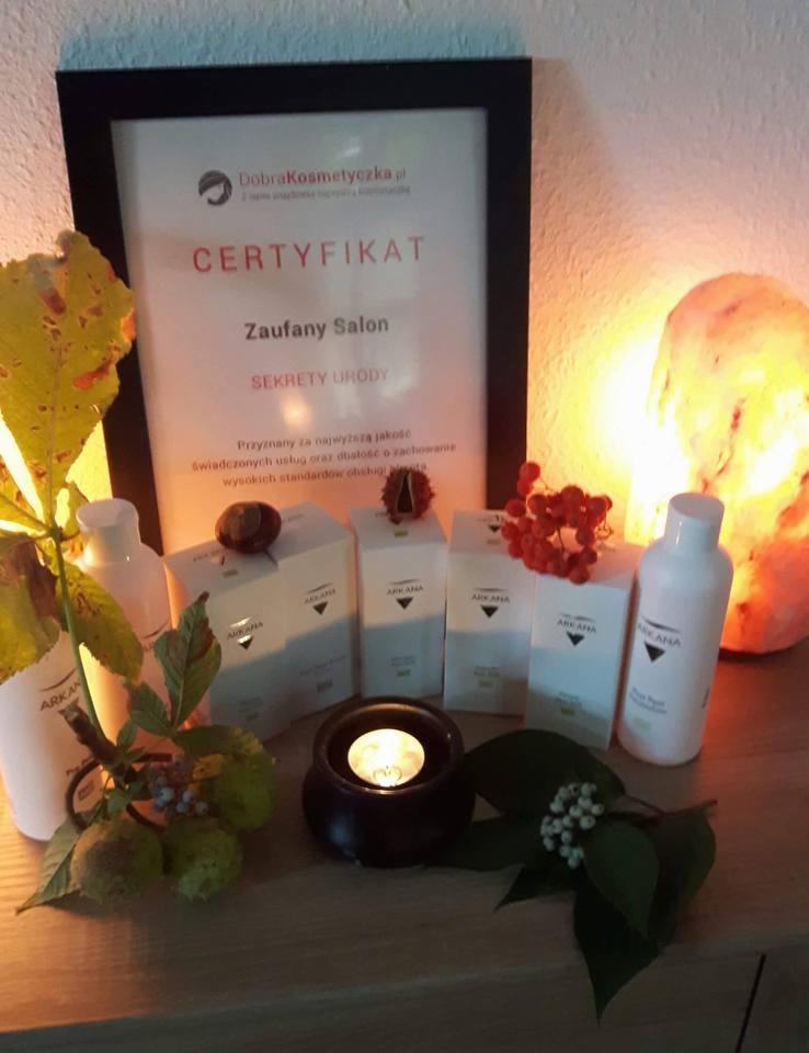 Certyfikat 'Zaufany Salon' DobraKosmetyczka.pl w ramce, otoczony kosmetykami Arkana, kasztanami, jarzębiną, świecą i lampą solną, tworzący klimatyczną kompozycję.
