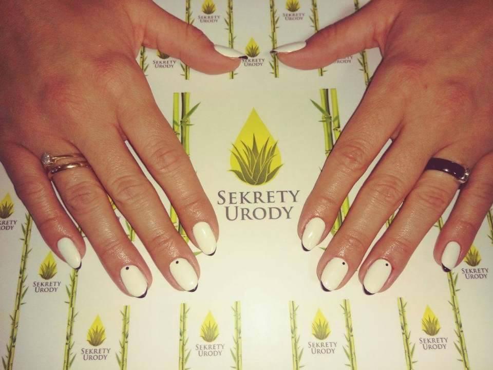 Dłonie z białym manicure hybrydowym z czarnymi końcówkami i drobnymi czarnymi kropeczkami na tle papieru firmowego salonu w Lublinie.