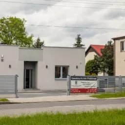 Nowoczesny budynek kancelarii prawno-podatkowej z szarymi panelami, widoczny szyld reklamowy z danymi kontaktowymi, otoczony zielenią i metalowym ogrodzeniem.