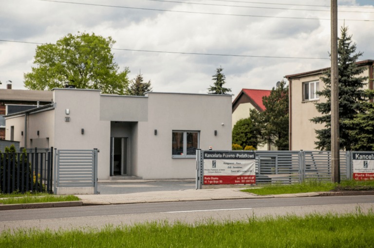 Nowoczesny budynek kancelarii prawno-podatkowej z szarymi panelami, widoczny szyld reklamowy z danymi kontaktowymi, otoczony zielenią i metalowym ogrodzeniem.