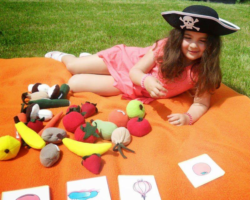 Dziewczynka w kapeluszu pirata leży na pomarańczowym kocu na trawie, otoczona pluszowymi warzywami i owocami oraz kartami z ich rysunkami.
