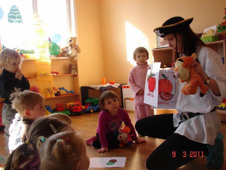 Nauczycielka przebrana za pirata, z pudełkiem z obrazkami owoców i pluszowym tygrysem, prowadzi zajęcia z grupą dzieci w przedszkolu, na podłodze leżą rysunki.