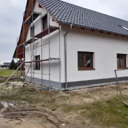 Świeżo otynkowany dom jednorodzinny z szarą podmurówką, widoczne rusztowania i teren po budowie.