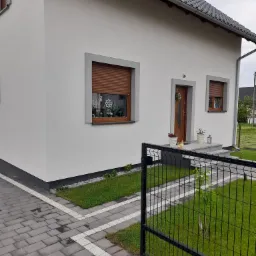 Biała elewacja domu jednorodzinnego z szarą stolarką okienną i brązowymi roletami, widok z zewnątrz przez czarne ogrodzenie panelowe na zadbany trawnik i kostkę brukową.