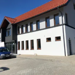 Nowoczesny budynek biurowy z brązowymi ramami okiennymi i czerwoną dachówką, z zaparkowanym niebieskim vanem przed wejściem