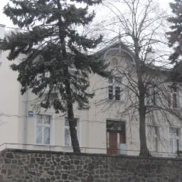 Elewacja dwupiętrowego domu z jasną fasadą, widoczna zza kamiennego murku, z numerem domu na ścianie i drzewami na pierwszym planie.