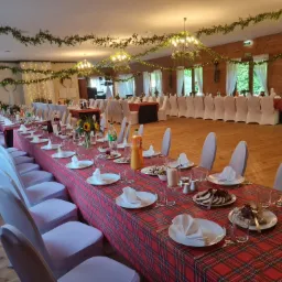 Długi stół z czerwonym obrusem w kratę, nakryty białymi talerzami i serwetkami, dekorowany kwiatami i girlandami świetlnymi, ustawiony w przestronnej sali z drewnianą podłogą i białymi krzesłami...