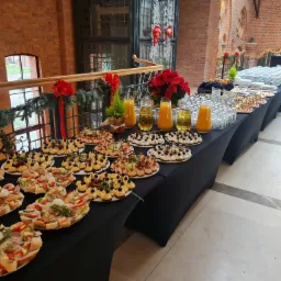 Długi stół cateringowy z czarnym obrusem w przestronnym wnętrzu z ceglanymi ścianami, na stole talerze z kanapkami, przekąskami, dzbanki z sokiem pomarańczowym i stosy szklanek, dekoracje...