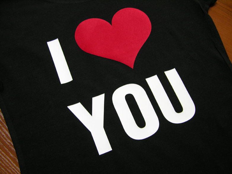 Czarna koszulka z białym napisem 'I YOU' i czerwonym sercem zamiast 'love', leżąca na drewnianym podłożu.