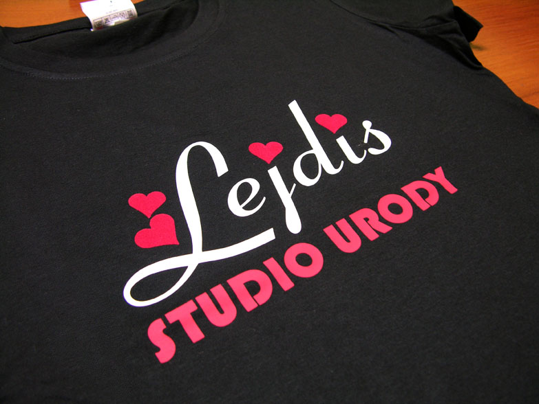 Czarna koszulka z białym napisem 'Lejdis' ozdobionym czerwonymi sercami i czerwonym napisem 'STUDIO URODY' poniżej, leżąca na drewnianym blacie.