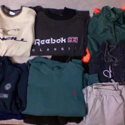 Stos pogniecionych ubrań sportowych używanych, w tym bluza Reebok z flagą brytyjską, kurtka dżinsowa i spodnie dresowe, ułożonych na szarym tle.