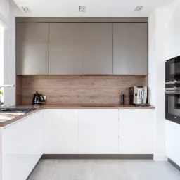 Nowoczesna kuchnia z białymi, błyszczącymi frontami szafek, drewnianym blatem i szarymi szafkami wiszącymi. Na blacie czajnik elektryczny i ekspres do kawy, obok zlewozmywaka koszyk z owocami...