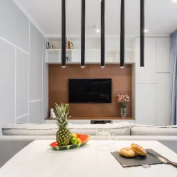 Jasne wnętrze salonu z białym stołem, owocami i pieczywem na pierwszym planie, telewizor na drewnianej ścianie w tle, minimalistyczne lampy wiszące i szare ściany z białymi geometrycznymi zdobieniami.