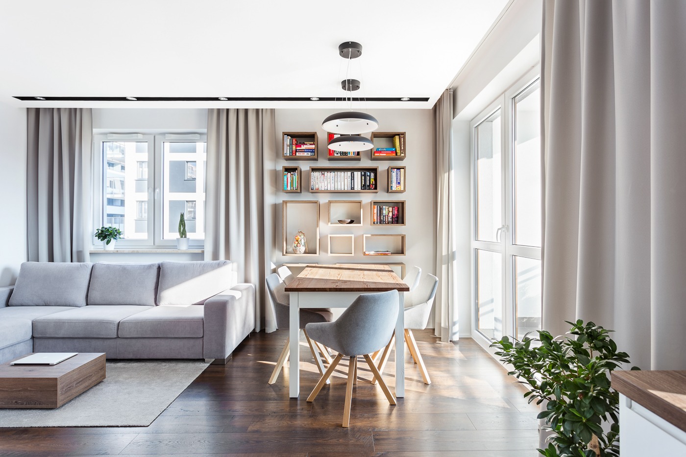 Jasne, nowoczesne wnętrze mieszkania z drewnianą podłogą, szarą sofą narożną, drewnianym stolikiem kawowym, stołem z krzesłami i półkami na książki na ścianie.