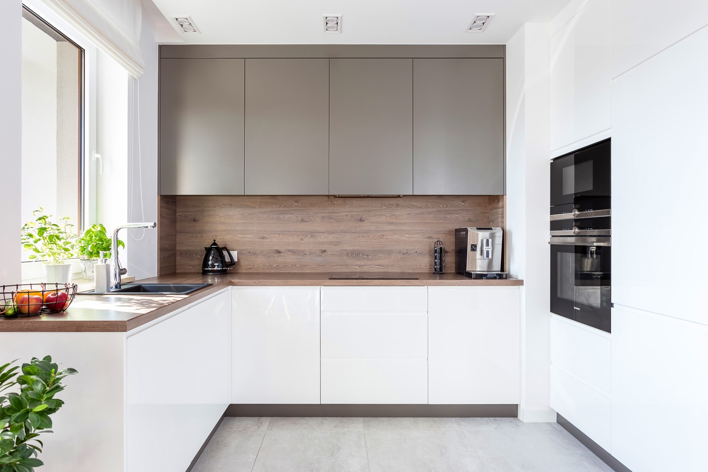 Nowoczesna kuchnia z białymi, błyszczącymi frontami szafek, drewnianym blatem i szarymi szafkami wiszącymi. Na blacie czajnik elektryczny i ekspres do kawy, obok zlewozmywaka koszyk z owocami...