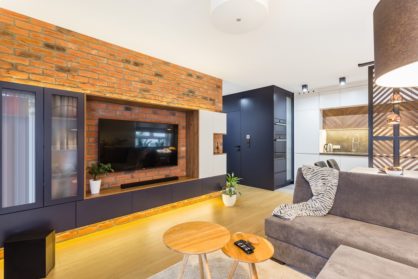 Nowoczesny salon z ceglaną ścianą, telewizorem, drewnianymi stolikami kawowymi i szarą sofą z kocem, oświetlenie LED pod szafkami RTV, widok na kuchnię w tle.
