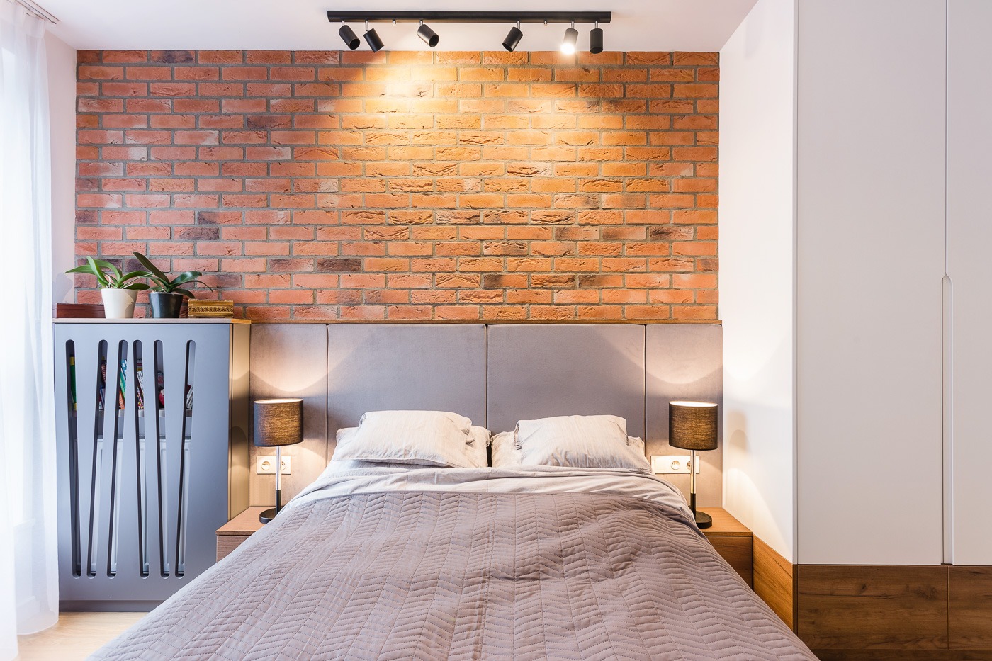 Sypialnia z ceglaną ścianą, szarym zagłówkiem, lampkami nocnymi z abażurami i grzejnikiem z ażurową obudową, oświetlona reflektorami sufitowymi.
