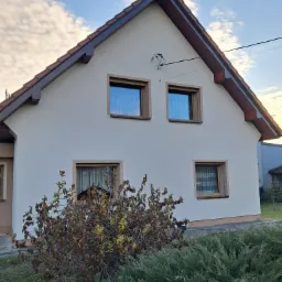 Dom jednorodzinny z jasną elewacją, brązowymi ramami okiennymi i dachem z czerwonej dachówki, widoczny krzew na pierwszym planie.